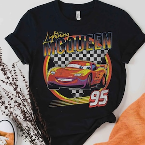 May include: Black t-shirt featuring a graphic of Lightning McQueen from the movie Cars. The design includes the text "Lightning McQueen" and the number "95" in a retro style, with a checkered flag background.