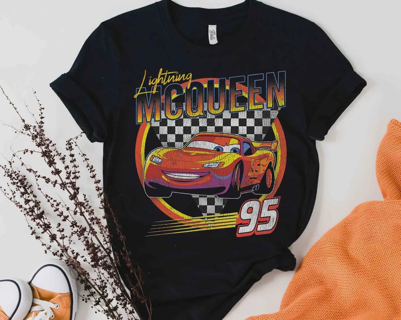 May include: Black t-shirt featuring a graphic of Lightning McQueen from the movie Cars. The design includes the text "Lightning McQueen" and the number "95" in a retro style, with a checkered flag background.
