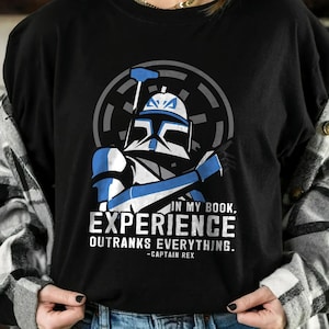 May include: Black t-shirt featuring a blue and white illustration of a Star Wars character, with the text "IN MY BOOK, EXPERIENCE OUTRANKS EVERYTHING. -CAPTAIN REX". The design is centered on the shirt.
