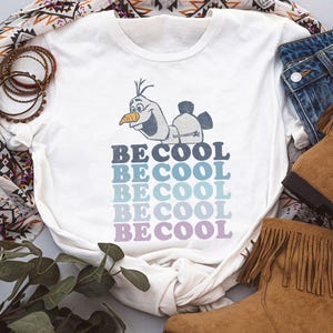 Puede incluir: Camiseta blanca con un personaje de dibujos animados y el texto "BE COOL" en degradado de azul y morado. La camiseta se combina con vaqueros y botas marrones con flecos, creando un look casual.