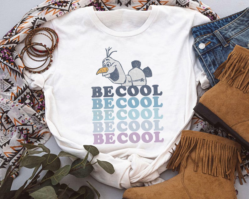 Puede incluir: Camiseta blanca con un personaje de dibujos animados y el texto "BE COOL" en degradado de azul y morado. La camiseta se combina con vaqueros y botas marrones con flecos, creando un look casual.