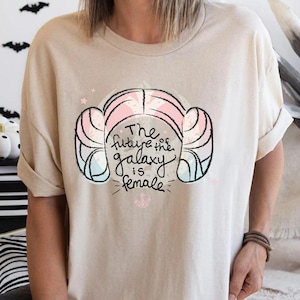 Puede incluir: Camiseta beige con un diseño de peinado y el texto "The future is female". El diseño incluye elementos rosas, azules y negros. La camiseta tiene mangas cortas y cuello redondo.