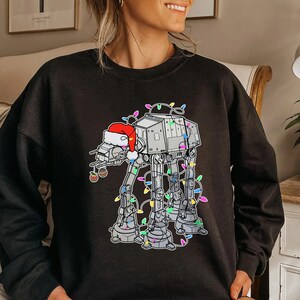 May include: Black sweatshirt featuring a festive design of an AT-AT walker wearing a Santa hat and adorned with colorful Christmas lights. A holiday ornament hangs from the walker's side. The design is a playful blend of Star Wars and Christmas themes.