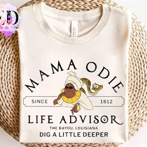 May include: Cream-colored t-shirt featuring a cartoon illustration of Mama Odie, a character with a turban and sunglasses, and a snake. The text reads "MAMA ODIE LIFE ADVISOR" with "SINCE 1812" and "DIG A LITTLE DEEPER".