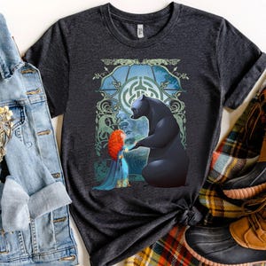 Puede incluir: Camiseta gris oscuro con un gráfico de una mujer pelirroja y un oso negro en una escena de bosque. El diseño incluye un fondo azul y verde con un patrón de laberinto. La camiseta está combinada con una chaqueta vaquera y botas.