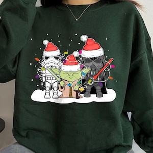 May include: Dark green sweatshirt featuring a festive design with Star Wars characters. Stormtrooper, Yoda, and Darth Vader are wearing Santa hats and Christmas lights. The design is on a white snow background.