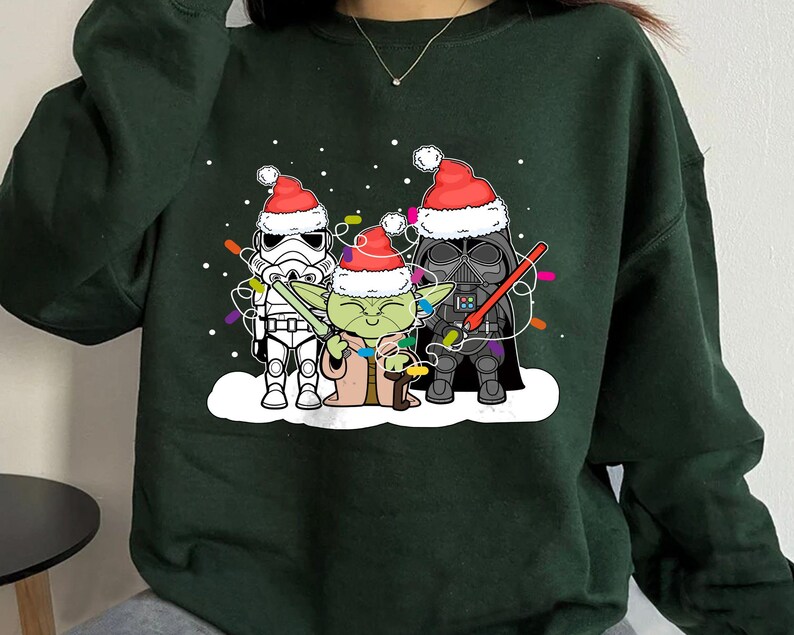 May include: Dark green sweatshirt featuring a festive design with Star Wars characters. Stormtrooper, Yoda, and Darth Vader are wearing Santa hats and Christmas lights. The design is on a white snow background.