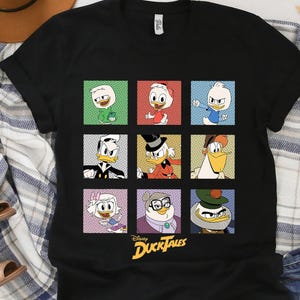 Op de afbeelding: Zwart T-shirt met een raster van negen cartoonkarakterportretten uit Disney's DuckTales. Elk personage staat in een vierkant met een verschillend gekleurde achtergrond. Het DuckTales-logo staat onderaan.