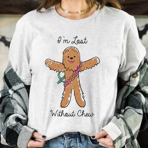 May include: A light grey t-shirt with a cartoon illustration of a brown, furry character wearing a pink and green bandolier. The text "I'm Lost Without Chew" is printed in black script below the character.