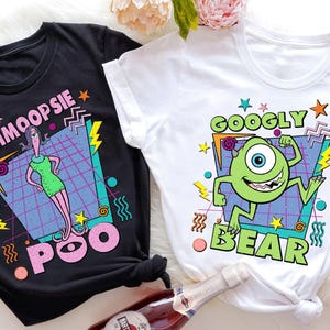 May include: Two graphic t-shirts, one black and one white. The black shirt features a cartoon woman with the text "SCHMOOPSIE POO". The white shirt displays a green monster with the text "GOOGLY BEAR". Both shirts have a retro 80s design.
