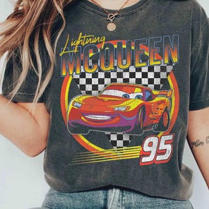 May include: Dark gray t-shirt featuring a graphic of Lightning McQueen from the movie Cars. The design includes the text "Lightning McQueen" in yellow and orange, the number "95" in red, and a checkered flag background.