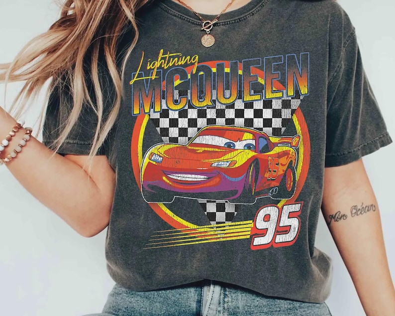 May include: Dark gray t-shirt featuring a graphic of Lightning McQueen from the movie Cars. The design includes the text "Lightning McQueen" in yellow and orange, the number "95" in red, and a checkered flag background.