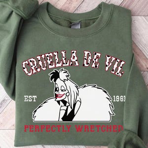 Pu&ograve; includere: Una felpa verde oliva con una grafica di Crudelia De Mon e la scritta "CRUELLA DE VIL" sopra la sua immagine. Il testo "PERFECTLY WRETCHED" &egrave; sotto l'immagine in rosso. L'anno "1961" &egrave; stampato anche sulla felpa.