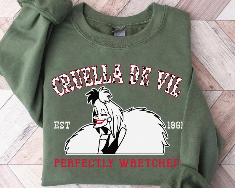 Pu&ograve; includere: Una felpa verde oliva con una grafica di Crudelia De Mon e la scritta "CRUELLA DE VIL" sopra la sua immagine. Il testo "PERFECTLY WRETCHED" &egrave; sotto l'immagine in rosso. L'anno "1961" &egrave; stampato anche sulla felpa.