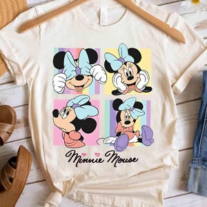 Pu&ograve; includere: T-shirt color crema con quattro pannelli con illustrazioni di Minnie Mouse. Ogni pannello mostra una posa diversa di Minnie, con un fiocco azzurro chiaro e sfondi colorati. Le parole "Minnie Mouse" sono scritte in corsivo sotto.