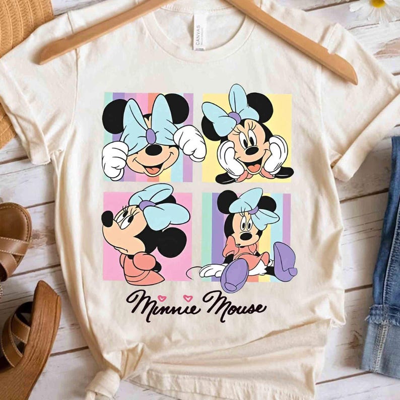 Pu&ograve; includere: T-shirt color crema con quattro pannelli con illustrazioni di Minnie Mouse. Ogni pannello mostra una posa diversa di Minnie, con un fiocco azzurro chiaro e sfondi colorati. Le parole "Minnie Mouse" sono scritte in corsivo sotto.