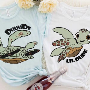 May include: Two t-shirts, one light blue and one white, each featuring a cartoon turtle graphic. The light blue shirt says "DUUUDE" above the turtle. The white shirt says "LIL DUDE" below the turtle. Both shirts are casual wear.