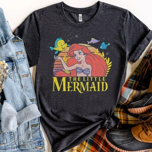 May include: Dark gray t-shirt featuring a graphic of Ariel from The Little Mermaid, with Flounder and other sea creatures. The text "THE LITTLE MERMAID" is in yellow. The shirt is styled with a denim jacket and brown boots.