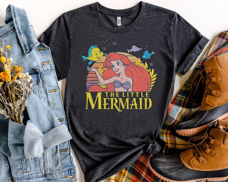 May include: Dark gray t-shirt featuring a graphic of Ariel from The Little Mermaid, with Flounder and other sea creatures. The text "THE LITTLE MERMAID" is in yellow. The shirt is styled with a denim jacket and brown boots.