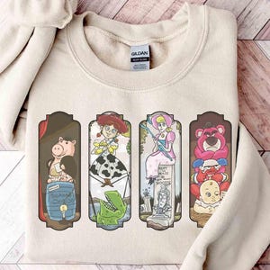 Puede incluir: Una sudadera beige con cuatro paneles rectangulares con personajes de dibujos animados. Los paneles muestran un cerdo, una vaquera, una mujer con un vestido azul y un oso rosa con un bebé. La sudadera tiene cuello redondo y mangas largas.