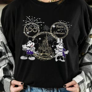 May include: Black sweatshirt featuring Mickey and Minnie Mouse with a castle graphic. The design includes the text "100 Years of Wonder" and fireworks. Mickey Mouse is wearing a purple outfit, and Minnie Mouse is wearing a purple bow.