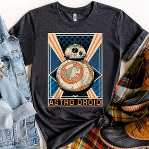 Puede incluir: Camiseta gris oscuro con un gráfico retro de un Astro Droid. El diseño presenta una ilustración detallada del droide con acentos naranjas y blancos, sobre un fondo geométrico azul y naranja. Las palabras "ASTRO DROID" están impresas debajo.