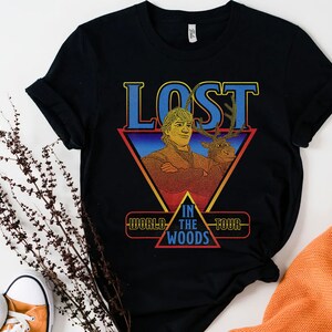 May include: A black short-sleeved t-shirt featuring a graphic print of a man and a reindeer. The text reads LOST IN THE WOODS WORLD TOUR. The design is set within a red and yellow triangle with a blue gradient background and bold blue lettering.