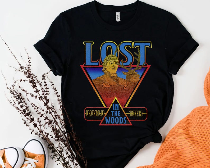 May include: A black short-sleeved t-shirt featuring a graphic print of a man and a reindeer. The text reads LOST IN THE WOODS WORLD TOUR. The design is set within a red and yellow triangle with a blue gradient background and bold blue lettering.
