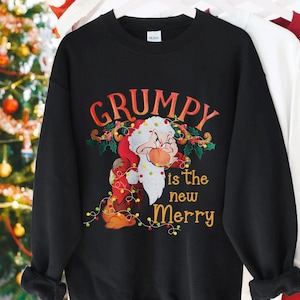 Peut inclure: Sweat-shirt noir avec l'inscription "GRUMPY is the new Merry" et un dessin de Grumpy de Blanche-Neige déguisé en Père Noël. Le motif comprend des guirlandes lumineuses et du houx.