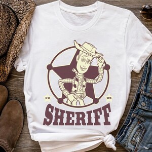Puede incluir: Camiseta blanca con un dise&ntilde;o de sheriff de dibujos animados dentro de una estrella. El dise&ntilde;o incluye la palabra "SHERIFF" en una fuente de estilo occidental, con los n&uacute;meros "19" y "95" a cada lado del emblema.