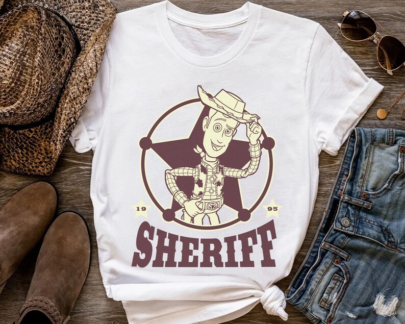 Puede incluir: Camiseta blanca con un dise&ntilde;o de sheriff de dibujos animados dentro de una estrella. El dise&ntilde;o incluye la palabra "SHERIFF" en una fuente de estilo occidental, con los n&uacute;meros "19" y "95" a cada lado del emblema.