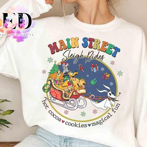 May include: White crewneck sweatshirt with a colorful graphic featuring cartoon dogs in a sleigh with the text "Main Street Sleigh Rides". The design includes a Christmas tree, gifts, and the words "hot cocoa, cookies, magical fun".