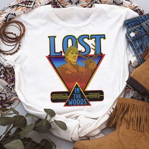 May include: A white short-sleeved t-shirt featuring a graphic print of a man and a reindeer inside a triangle. The text reads LOST WORLD IN THE WOODS TOUR. The design uses blue, red, and yellow colors with a retro aesthetic and a crew neckline.