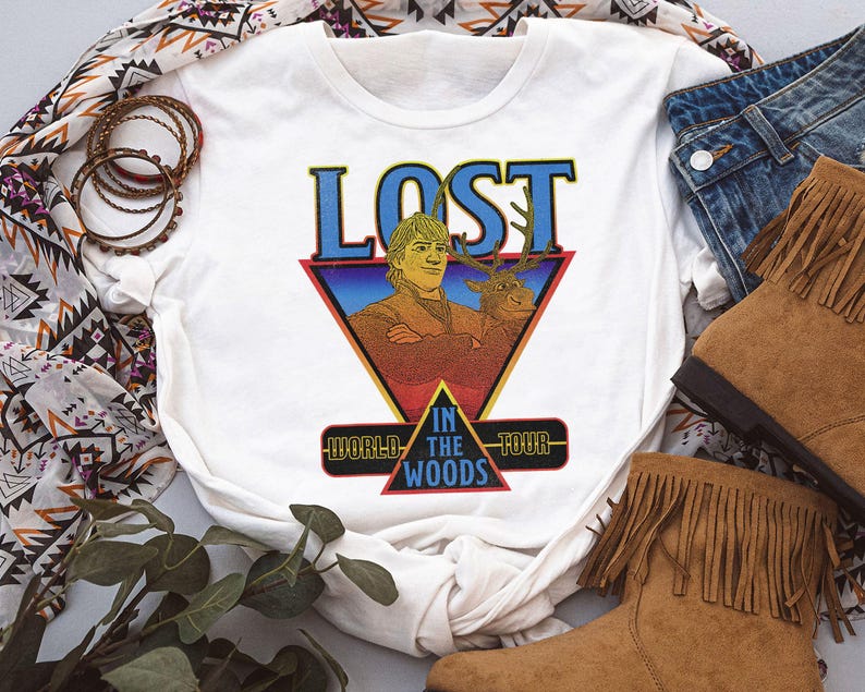 May include: A white short-sleeved t-shirt featuring a graphic print of a man and a reindeer inside a triangle. The text reads LOST WORLD IN THE WOODS TOUR. The design uses blue, red, and yellow colors with a retro aesthetic and a crew neckline.