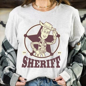 Puede incluir: Camiseta gris claro con un dibujo de sheriff de dibujos animados dentro de un c&iacute;rculo y una estrella. La palabra "SHERIFF" est&aacute; impresa debajo en granate, con los n&uacute;meros "19" y "95" a cada lado. Tema cl&aacute;sico del oeste.