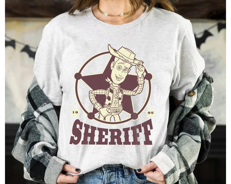 Puede incluir: Camiseta gris claro con un dibujo de sheriff de dibujos animados dentro de un c&iacute;rculo y una estrella. La palabra "SHERIFF" est&aacute; impresa debajo en granate, con los n&uacute;meros "19" y "95" a cada lado. Tema cl&aacute;sico del oeste.