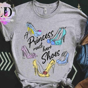 May include: A heather gray t-shirt featuring a graphic print of five colorful high-heeled shoes and the black text A Princess must have Shoes. The shoes are illustrated in blue, pink, and yellow with names written on each individual shoe.