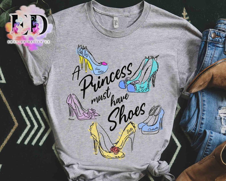 May include: A heather gray t-shirt featuring a graphic print of five colorful high-heeled shoes and the black text A Princess must have Shoes. The shoes are illustrated in blue, pink, and yellow with names written on each individual shoe.