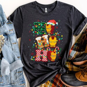 May include: Dark gray t-shirt featuring a festive design with the words "HO HO HO" in gingerbread cookie style, with Iron Man helmet graphics. The design includes Christmas lights and a Santa hat. The shirt is displayed with a denim jacket and boots.