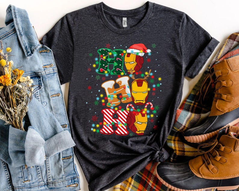 May include: Dark gray t-shirt featuring a festive design with the words "HO HO HO" in gingerbread cookie style, with Iron Man helmet graphics. The design includes Christmas lights and a Santa hat. The shirt is displayed with a denim jacket and boots.