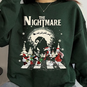 Pode incluir: Camisola verde escuro com um gr&aacute;fico de "O Estranho Mundo de Jack". O design apresenta personagens atravessando uma faixa de pedestres, uma lua assustadora e as palavras "The Nightmare" em branco. Flocos de neve e &aacute;rvores de Natal tamb&eacute;m s&atilde;o vis&iacute;veis.