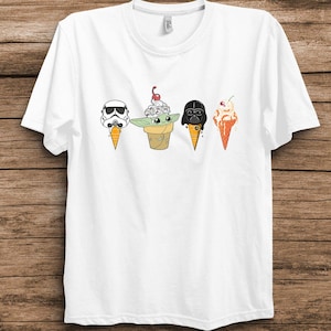 Puede incluir: Camiseta blanca con cuatro conos de helado de dibujos animados que representan personajes de Star Wars. Los personajes incluyen un Stormtrooper, Baby Yoda, Darth Vader y un personaje con un cono naranja y una cereza. La camiseta está sobre un fondo de madera.