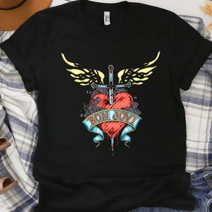 Puede incluir: Camiseta negra con un diseño gráfico que presenta un corazón rojo atravesado por una espada, adornado con alas amarillas y una pancarta que dice "BON JOVI". El diseño es detallado y colorido, con una estética vintage.