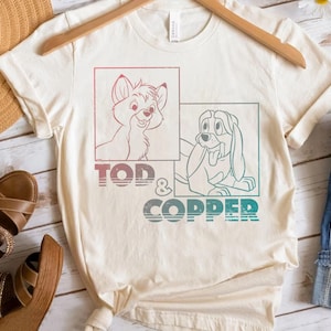 May include: Cream-coloured t-shirt featuring a graphic of Tod and Copper, with the text "TOD & COPPER" in a gradient of red, blue, and green. The shirt is displayed on a wooden hanger.