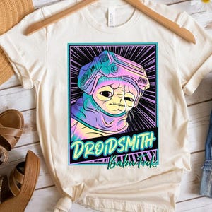 May include: A cream-coloured t-shirt with a vibrant graphic of a creature wearing a helmet. The text "DROIDSMITH" is displayed in a retro font, with "Baby Pink" below. The design has a retro, neon aesthetic.
