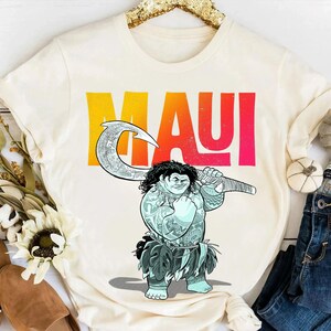 May include: Cream-colored t-shirt featuring a cartoon illustration of Maui holding a large hook. The word "MAUI" is printed in large, colorful letters across the top. The shirt is displayed with jeans and decorative items.