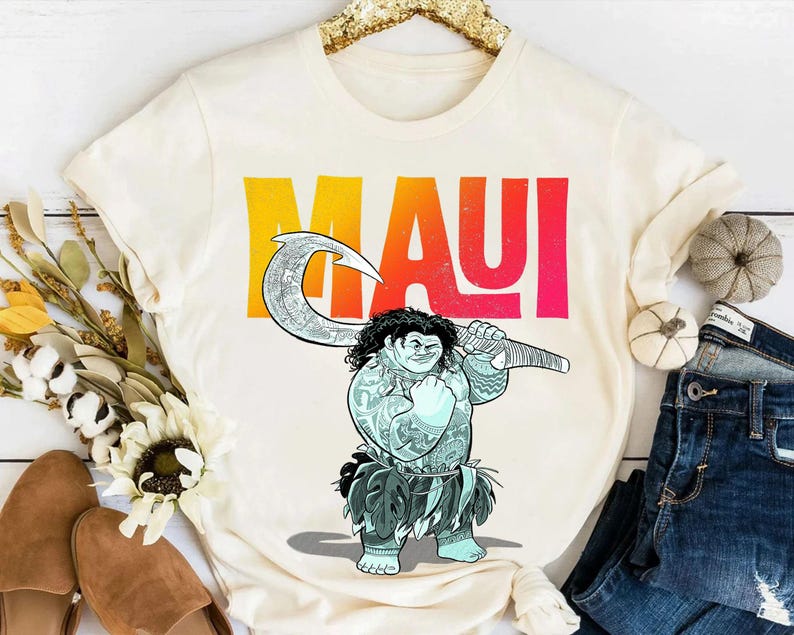 May include: Cream-colored t-shirt featuring a cartoon illustration of Maui holding a large hook. The word "MAUI" is printed in large, colorful letters across the top. The shirt is displayed with jeans and decorative items.