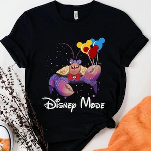 May include: Black t-shirt featuring a cartoon crab character holding colorful balloons. The crab has purple claws and a red bow tie. The words "Disney Mode" are written in white script below the crab.