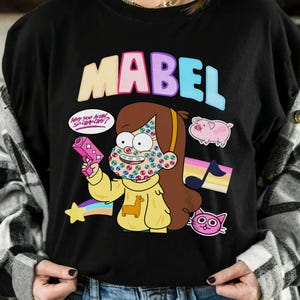 Puede incluir: Camiseta negra con el nombre "MABEL" en letras coloridas. El gráfico incluye un personaje de dibujos animados con detalles faciales coloridos, sosteniendo una pistola de juguete rosa y rodeado de otras imágenes de dibujos animados.