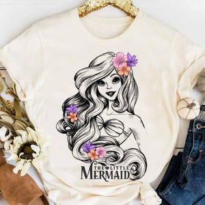 May include: Cream-colored t-shirt featuring a black and white illustration of a mermaid with long flowing hair adorned with purple and orange flowers. The text "THE LITTLE MERMAID" is printed below the illustration.